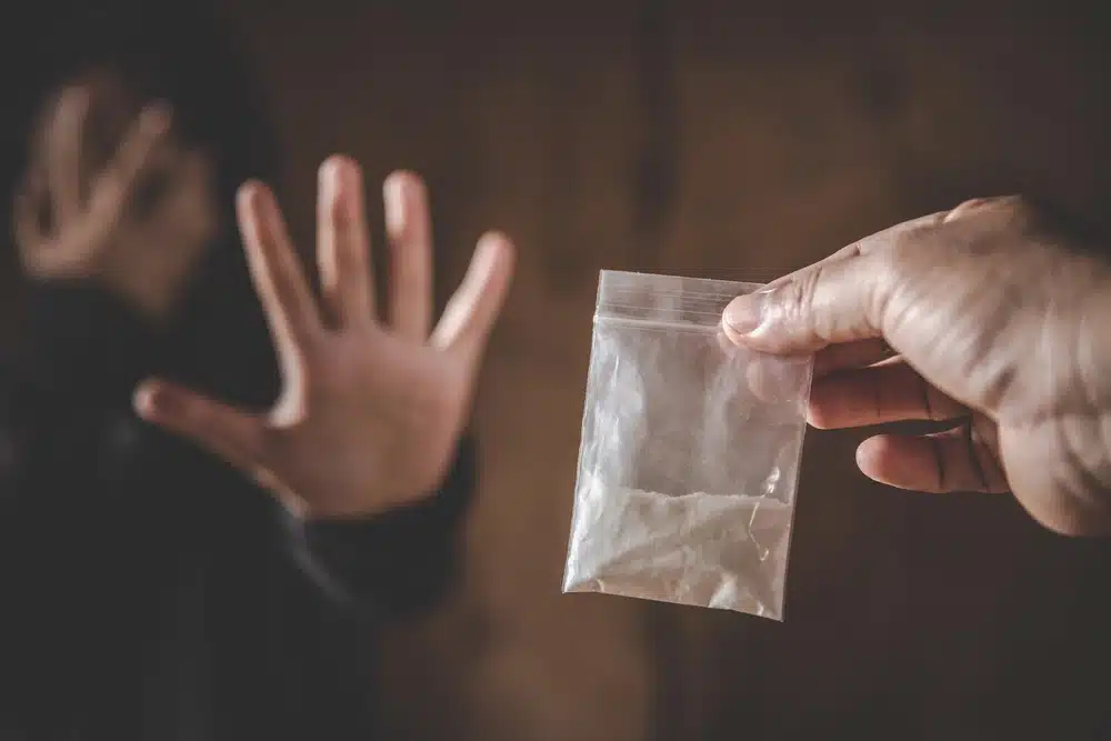 A person in a hooded sweatshirt raising their hand in refusal as another person offers a small plastic bag of white powder