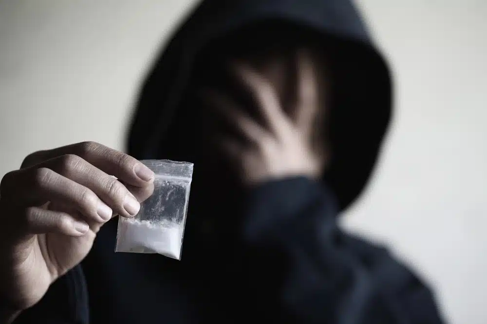 A person in a hooded sweatshirt holding a small plastic bag of white powder while covering their face with their other hand.