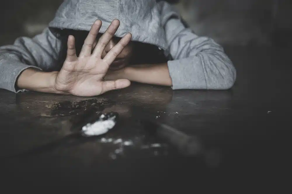  person in a gray hoodie lying on a table with scattered drug paraphernalia, raising their hand to refuse or shield themselves